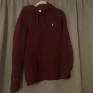 Vinröd hoodie från Lyle & Scott - Säljer en snygg vinröd hoodie från Lyle & Scott med en gul logga på bröstet. Tröjan har en klassisk design med huva och dragsko samt en praktisk magficka. Perfekt för kyliga dagar!