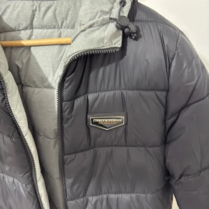Svart och grå vändbar pufferjacka från Superdry - Snygg vändbar pufferjacka från Superdry i svart och grått. Jackan har en praktisk dragkedja och justerbar huva. Perfekt för kyliga dagar med sin quiltade design som håller värmen. Två sidofickor för extra förvaring.