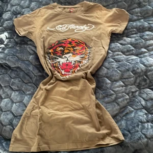 Brun t-shirt från Ed Hardy - Snygg brun t-shirt/kort klänning/lång top från Ed Hardy med ett stort tigertryck i rött, svart och orange på framsidan. T-shirten har korta ärmar och ett coolt, grafiskt mönster. Perfekt för dig som gillar att sticka ut!