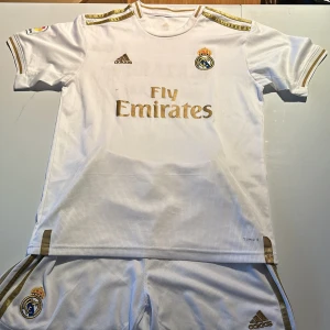 Vit Real Madrid fotbollströja och shorts med guldtryck - Snygg vit Real Madrid fotbollströja med guldiga detaljer och tryck. Tröjan har korta ärmar och klubbens emblem på bröstet. Nummer 23 och namnet Hazard pryder baksidan. Perfekt för fans av laget!