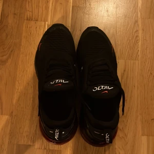Nike Air Max 270 i svart och röd - Snygga Nike Air Max 270 sneakers i svart med röda detaljer. Skorna har en synlig Air Max-enhet i hälen och klassisk snörning. Perfekta för en sportig look. Dock är skorna svlivit slipade i backsidan. Luften är ochså slut o bublorna men det går att pumpa😃