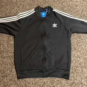 Svart träningsjacka från Adidas - Säljer en klassisk svart träningsjacka från Adidas med de ikoniska tre ränderna längs ärmarna. Jackan har dragkedja framtill och Adidas-loggan broderad på bröstet. Perfekt för en sportig och retro look! Den har storlek L men är typ en M.