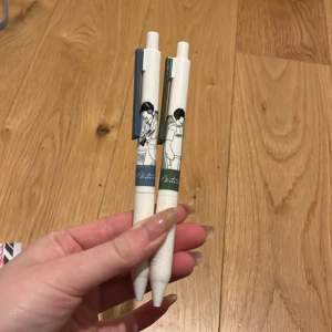 Vita pennor med illustrationer - Två vita pennor med unika illustrationer av personer. Pennorna har en blå och en grön detalj nära toppen och en tryckmekanism för att fälla ut spetsen. Perfekta för att ge skrivandet en personlig touch.