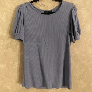 Blå ribbad topp från KappAhl - Snygg blå ribbad topp från KappAhl med korta puffärmar. Perfekt för en stilren look. Toppen har en rund halsringning och är tillverkad i ett mjukt material.