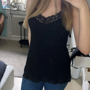 Svart linne med spetsdetaljer från Vero Moda - Snyggt svart linne från Vero Moda med spetsdetaljer vid halsringningen och nederkanten. Perfekt för en stilren look. Justerbara axelband för bästa passform. Bra kvalité 😊