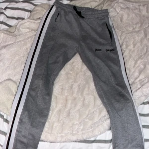 Grå träningsset från Palm Angels - Snyggt grått träningsset från Palm Angels med svarta och vita ränder längs ärmar och ben. Tröjan har dragkedja och ribbade muddar, medan byxorna har en bekväm resår i midjan. Perfekt för en sportig look.