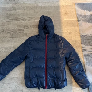 Mörkblå pufferjacka med röd dragkedja - Säljer en snygg mörkblå pufferjacka med en iögonfallande röd dragkedja. Jackan har en huva och är perfekt för kyligare dagar. Den quiltade designen ger extra värme och stil.