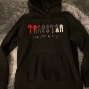Svart hoodie från Trapstar  - Snygg svart hoodie från Trapstar med färgglad text på bröstet i rött, vitt och grått. Perfekt för en casual look med sin bekväma passform och klassiska design. Huvan har dragsko för justering.