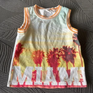 Färgglad linnetopp med Miami-tryck - Säljer en färgglad linnetopp med tryck av palmer och texten 'MIAMI' i stora bokstäver. Toppen har orange detaljer vid ärmhålen och halsen, perfekt för sommardagar.