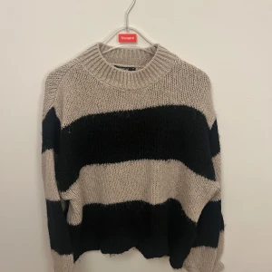 Randig stickad tröja från Gina  - Säljer en mysig stickad tröja från Gina  med breda ränder i beige och svart. Tröjan har en rund halsringning och långa ärmar, perfekt för kyliga dagar. Den är både stilren och bekväm.