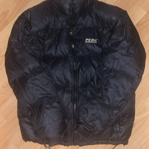 Svart dunjacka från Peak Performance - Säljer en svart dunjacka från Peak Performance. Jackan har en dragkedja framtill och är quiltad för extra värme. Perfekt för kyliga dagar. Har ett hål på höger arm som är lagat med en patch, sista bilden. Annars i använt skick 