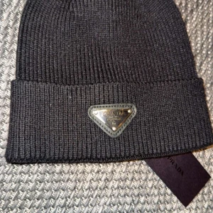 Prada mössa Svart/Grå - Snygg ribbad mössa från Prada i både svart och grått. Mössan har en uppvikt kant och en metalllogga framtill. Perfekt för att hålla värmen med stil.