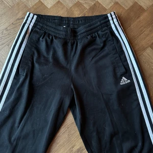 Svarta byxor från Adidas - Snygga svarta byxor från Adidas med de klassiska tre ränderna längs sidorna. Byxorna har en elastisk midja och är perfekta för sport och fritid. Adidas-loggan är diskret placerad på ena benet.