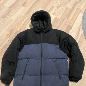 Svart och blå pufferjacka från H&M - Säljer en svart och blå pufferjacka från H&M. Jackan har en dragkedja framtill och två fickor med dragkedjor. Den är vattenavvisande och har en huva för extra skydd mot väder och vind. Storlek S men passar mer M