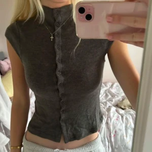 Grå ribbad topp - Säljer en snygg grå ribbad topp med knappar framtill. Toppen har en tight passform och korta ärmar, perfekt för en stilren look. Den är enkel att matcha med jeans eller kjol.