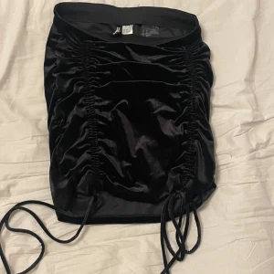 Svart sammetskjol från Divided - Snygg svart sammetskjol från Divided med snörning på sidorna. Kjolen har en tight passform och är perfekt för en trendig look. Den är i storlek XS och har en glansig finish som ger en lyxig känsla.