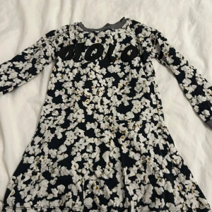 Popcorn klänning från Molo - Söt popcorn klänning från Molo med svart bas och vita popcorn. Klänningen har långa ärmar och ett bekvämt snitt, perfekt för lekfulla dagar. Texten 'MOLO' pryder framsidan för en extra cool touch. Klänningen kommer tvättas och strykas innan den skickas! ⭐️