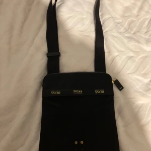 Svart axelväska från Boss - Snygg svart axelväska från Boss med justerbar rem och dragkedja upptill. Perfekt för att bära med sig det nödvändigaste med stil. Diskret och elegant design som passar till många tillfällen, saknar märke på främre sida .