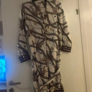 Mönstrad långklänning med knappar - Elegant långklänning med ett unikt mönster i beige, brunt och blått. Klänningen har långa ärmar och knappar framtill. Perfekt för en stilfull look.