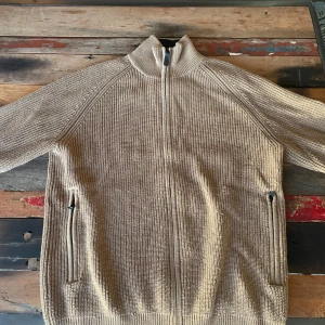 Beige stickad tröja från J.Lindeberg - Säljer en snygg beige stickad tröja från J.Lindeberg. Köpt för över 2000 kr på Stockholm Boulevard. Tröjan har en dragkedja framtill och en hög krage med kontrastfärg i svart. Perfekt för kyliga dagar! Skriv privat för fler bilder eller andra frågor! 