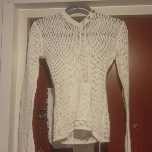 Vit stickad tröja med knappar - Säljer en vit stickad tröja med unika knapplösningar vid halsen. Tröjan har en djup v-ringning och långa ärmar och luva perfekt för lager-på-lager under kyliga dagar. Den är både stilren och bekväm, vilket gör den till ett mångsidigt tillskott i garderoben.