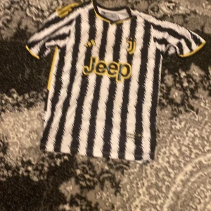 Juventus fotbollströja med Pogba 10 - Snygg Juventus fotbollströja från Adidas med Pogba och nummer 10 på ryggen. Tröjan är randig i svart och vit med gula detaljer och korta ärmar. Perfekt för alla Juve-fans!