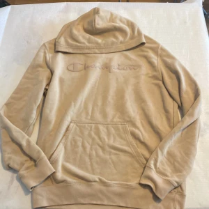 Beige hoodie från Champion - Säljer en beige hoodie från Champion med broderad logga på bröstet. Tröjan har en stor ficka framtill och långa ärmar. Perfekt för en avslappnad stil. Små hål på ryggen och vänstra axlen. 