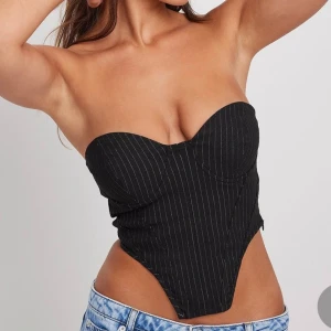 Svart randig korsett - Snygg svart korsett med tunna vita ränder. Den är axelbandslös och har en tight passform som framhäver figuren. Perfekt för en utekväll eller fest!