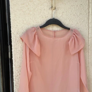 Blus - Fin blus i aprikos färg. Lång ärm. Knapp bak. Volang upptill på ärm. 