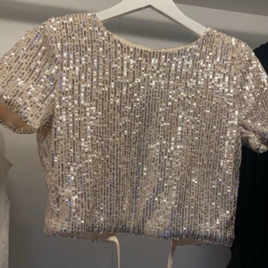 Glittrig beige paljettopp - Säljer en superfin glittrig topp. Den är kortärmad och croppad, perfekt för festliga tillfällen. Toppen har en liten öppning i ryggen som ger en extra snygg detalj. Passar perfekt till nyår eller andra festliga events! Man kan antigen ha med linne under eller utan😇 