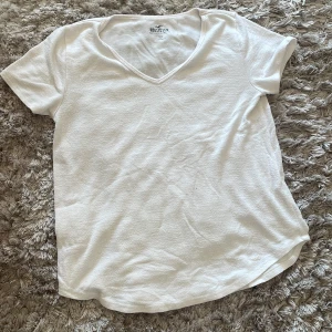 Vit t-shirt från Hollister - Säljer en enkel och stilren vit t-shirt från Hollister. Den har en v-ringad hals och korta ärmar, perfekt för en avslappnad look. Materialet känns mjukt och bekvämt, idealiskt för vardagsbruk.