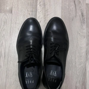 Svarta skinnskor från Pace - Eleganta svarta skinnskor från Pace med klassisk snörning. Perfekta för en stilren look. Skorna har en rundad tå och en låg klack, vilket ger en bekväm passform.