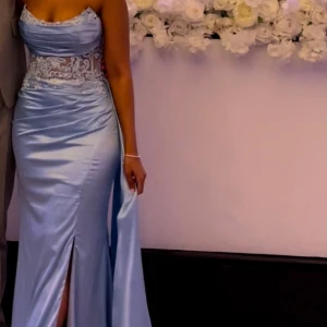 Ljusblå långklänning med spetsdetaljer - Klänningen användes endast för ett tillfälle och kemtvättades och strukits inför försäljning. (Är 160 cm, byst 75 C), men om man är mindre i bysten så kan man använda fashion tape. Klänningen kostade 3600 kr. Färgen är ljusblå men kan se något mörkare ut beroende på ljuset i vilket bilden tas. 