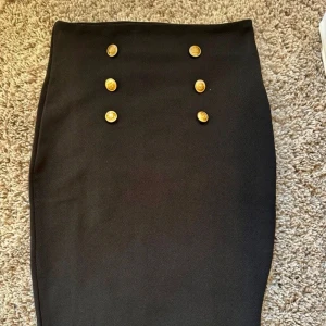 Pennkjol knälång Svart kjol med guldknappar - Elegant svart kjol med sex dekorativa guldknappar framtill. Kjolen har en klassisk och stilren design som passar perfekt för en chic look. Den är knälång och har en tight passform som framhäver figuren.