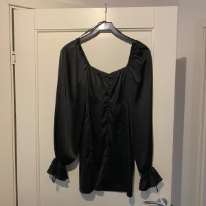 Svart långärmad klänning - Elegant svart klänning med långa ärmar och volangdetaljer vid ärmsluten. Klänningen har en v-ringad design och är tillverkad i ett glansigt material som ger en lyxig känsla. Perfekt för en stilren look.