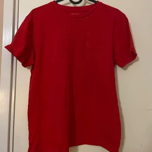 Röd t-shirt - Säljer en klassisk röd t-shirt. Den har en enkel design med korta ärmar och en liten bröstficka. Perfekt för en avslappnad stil. Tillverkad i mjukt material för bekväm passform.