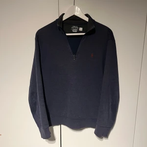 Mörkblå halvzip Ralph Lauren - Säljer en snygg mörkblå halvzip från Polo Ralph Lauren med dragkedja och broderad logga i rött på bröstet. Tröjan har en hög krage och långa ärmar, perfekt för kyligare dagar. Passar både till vardags och mer avslappnade tillfällen.