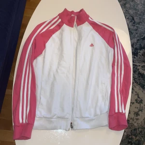 Vit och rosa träningsjacka från Adidas - Vit och rosa träningsjacka från Adidas med klassiska ränder längs ärmarna. Jackan har en dragkedja framtill och en hög krage. Den är märkt som storlek M, men eftersom den är lite kortare i ärmarna skulle jag lägga den mellan S-M. (Om man har +2 i apindex dvs)