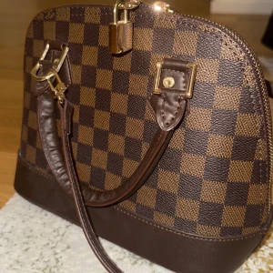 Brun rutig handväska/axelremsväska - Snygg brun handväska med klassiskt rutigt mönster och guldfärgade detaljer. Väskan har en dragkedja upptill och två handtag samt tillhörande axelband. Perfekt för att ge en stilren touch till din outfit.