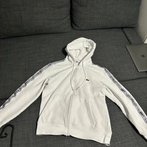 Vit hoodie från Lacoste - Säljer en stilren vit hoodie från Lacoste med dragkedja och klassisk passform. Den har  ärmar med märkets logotyp längs ärmarna och en liten krokodil på bröstet. Perfekt för en avslappnad stil.