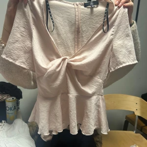 Rosa blus  - Säljer en söt rosa blus med korta ärmar och en knytning framtill. Blusen har en volangdetalj vid nederkanten och en dragkedja i ryggen. Perfekt för en somrig look!