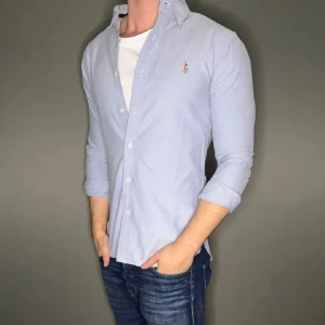 Ljusblå skjorta med broderad logga - Snygg ljusblå skjorta från Ralph Lauren med broderad logga på bröstet. Skjortan har långa ärmar och knappar framtill. Perfekt för en stilren look. Bra skick, pris kan diskuteras 