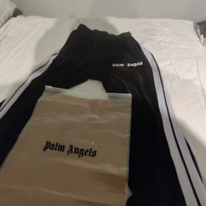 Svarta byxor från Palm Angels - Snygga svarta byxor från Palm Angels med vita sidoremsor. Byxorna har en elastisk midja och Palm Angels-loggan tryckt på framsidan. Perfekta för en avslappnad stil. Priset är inte hugget i sten.