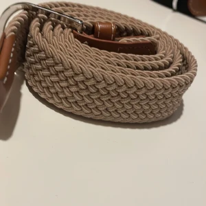  flätat bälte - Snyggt beige flätat bälte med bruna läderdetaljer och silverfärgat spänne. Perfekt för att ge en stilren touch till din outfit.