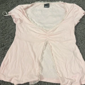 Rosa topp  - Söt ljusrosa topp från Gina Tricot med spetsdetaljer längs halsringningen och ärmslut. Toppen har en v-ringad design med rynkdetalj framtill och korta ärmar. Perfekt för en feminin look.