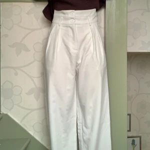 Vita högmidjade byxor - Snygga vita högmidjade byxor med vida ben och stilrena veck framtill. Byxorna har en klassisk design med knappar i midjan och passar perfekt för en elegant look. Aldrig använda