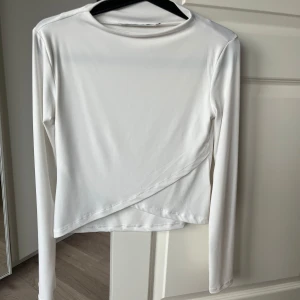 Vit långärmad topp - Säljer en stilren vit långärmad topp från Lager 157 i storlek XS. Toppen har en unik omlott-design framtill som ger en snygg detalj. Perfekt för att matcha med olika outfits. Två ”trådar” som ej syns på håll, inget man lägger märke till på! 