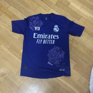 Real Madrid tröja PRIS KAN DISSKUTERAS - Snygg lila fotbollströja från Adidas med korta ärmar och coolt rosmönster. Tröjan har tryck med 'Emirates Fly Better' och Real Madrid-logga. Perfekt för fotbollsfans som vill sticka ut på planen!