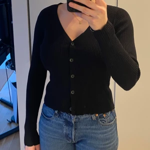 Svart ribbad kofta - Ssvart ribbad kofta med knappar framtill. Koftan har en v-ringad design och långa ärmar med små volang detaljer. 