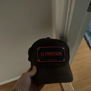 Svart keps från John Hatter & Co - Snygg svart keps med texten 'El Profesor' i rött framtill. Kepsen har en justerbar baksida för perfekt passform. Perfekt för en cool och avslappnad stil. Allt original ingår! Priset är inte hugget i sten. Den är rätt så brun pågrund av solen 